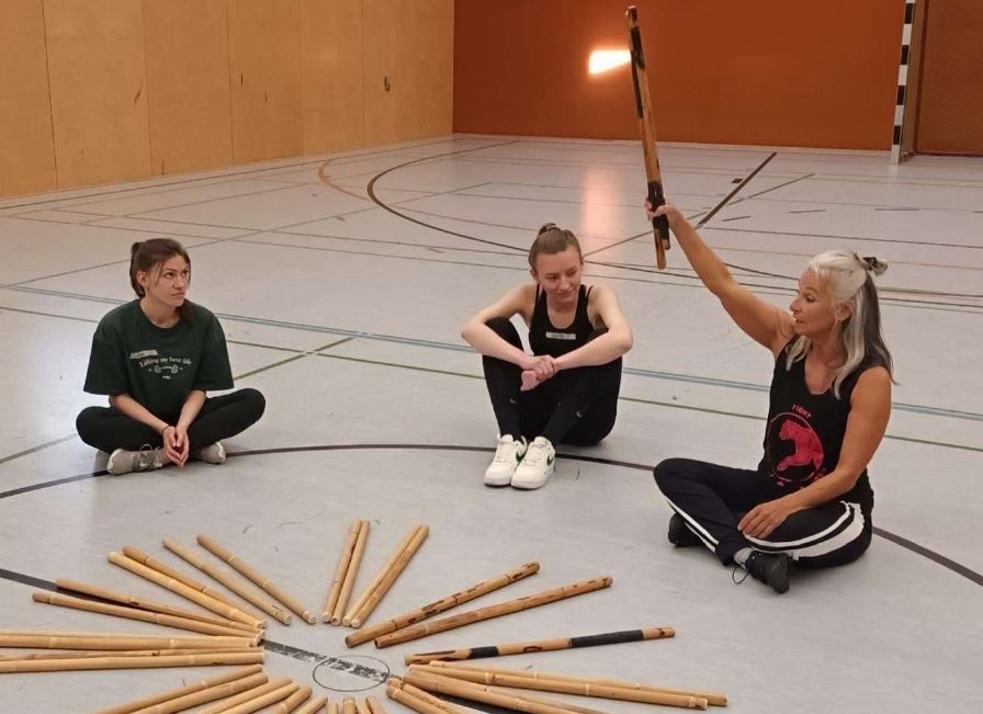 Eine Frau sitzt mit mehreren Mädchen in einer Sporthalle und hält einen Stock hoch.